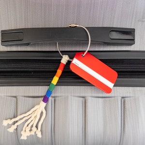 May include: A gray suitcase with a black handle and a red luggage tag attached to a rainbow-colored string. The tag has a white stripe across the middle.