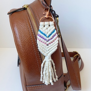 May include: A white macrame keychain with a pink, purple, and blue stripe pattern and a white tassel attached to a brown leather backpack.