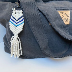 May include: A white and blue macrame keychain with a tassel hanging from a dark gray bag with a brown leather patch that says "Feel Good" with a mountain and trees graphic.