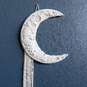 May include: A white macrame crescent moon wall hanging with a long fringe. The moon is made of white yarn and has a textured surface. The fringe is also made of white yarn and hangs down from the bottom of the moon.