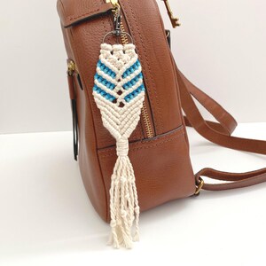 May include: A white macrame keychain with a blue and white geometric pattern. The keychain is attached to a brown leather backpack.