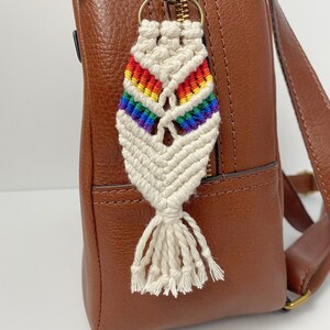 May include: A rainbow-colored macrame keychain with a white tassel attached to a gold key ring. The keychain is hanging from a brown leather backpack.