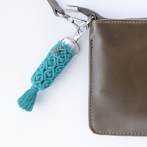 May include: Teal blue macrame keychain with a crescent moon charm attached to a silver key ring. The keychain is hanging from a brown leather purse.