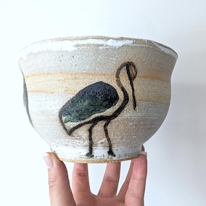 May include: A ceramic bowl with a textured, off-white finish and horizontal tan bands. The exterior features a hand-painted illustration of a bird with a dark green body and black outlines. The bowl has a wide, flared rim and a tapered base.