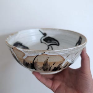 May include: A ceramic bowl with a white glazed interior featuring a black painted crane illustration. The exterior displays a natural tan clay base with white glaze drips and black brushstroke details around the lower portion of the vessel.