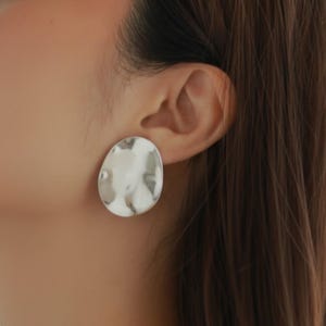 Puede incluir: Pendientes ovalados plateados con textura martillada. Los pendientes son grandes y tienen una superficie reflectante. También se ve un delicado collar de oro.