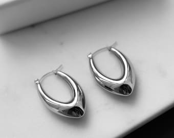 May include: A pair of silver hoop earrings with a unique oval shape. The earrings are on a white surface with a box that says "Rebel Rebel" in the background.