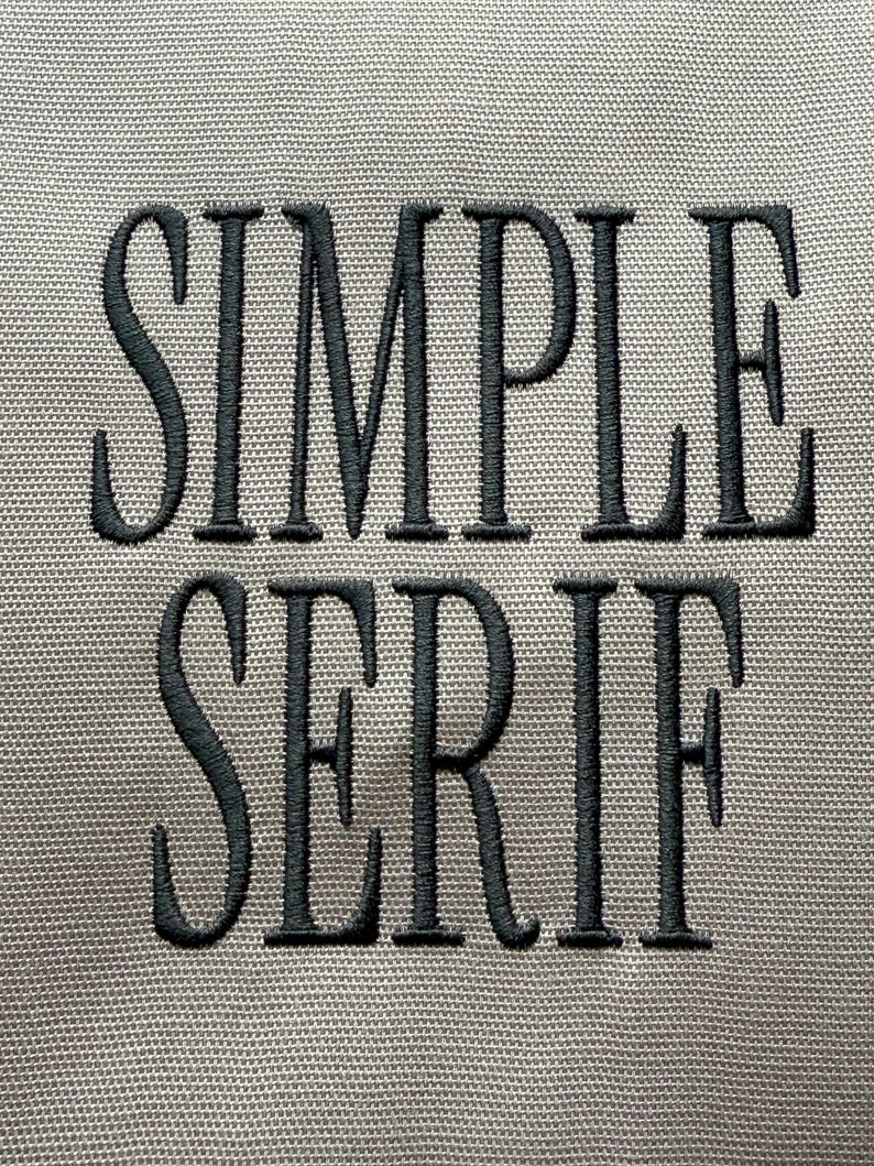 Op de afbeelding: Close-up van een grijze stof met de woorden "SIMPLE SERIF" geborduurd in donkergrijs garen. De tekst is vetgedrukt, met een schreefletter, en de stof heeft een subtiele geweven textuur.
