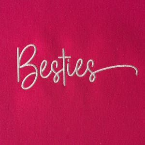 May include: A close-up of a vibrant fuchsia fabric with the word "Besties" embroidered in white cursive script. The text is centered and the script has a delicate, flowing style. The background is a solid, bright pink.