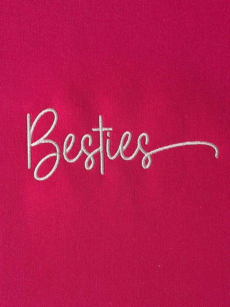 May include: A close-up of a vibrant fuchsia fabric with the word "Besties" embroidered in white cursive script. The text is centered and the script has a delicate, flowing style. The background is a solid, bright pink.
