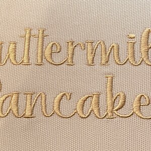 May include: A white kitchen towel with the words "Buttermilk Pancakes" embroidered in gold thread.