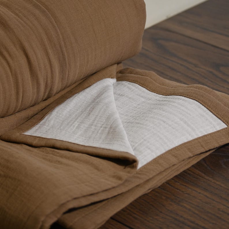 May include: A folded brown blanket with a white interior. The blanket is made of a soft, textured fabric. The edges are neatly finished with a contrasting brown trim. The blanket is partially rolled up, revealing its layered construction.