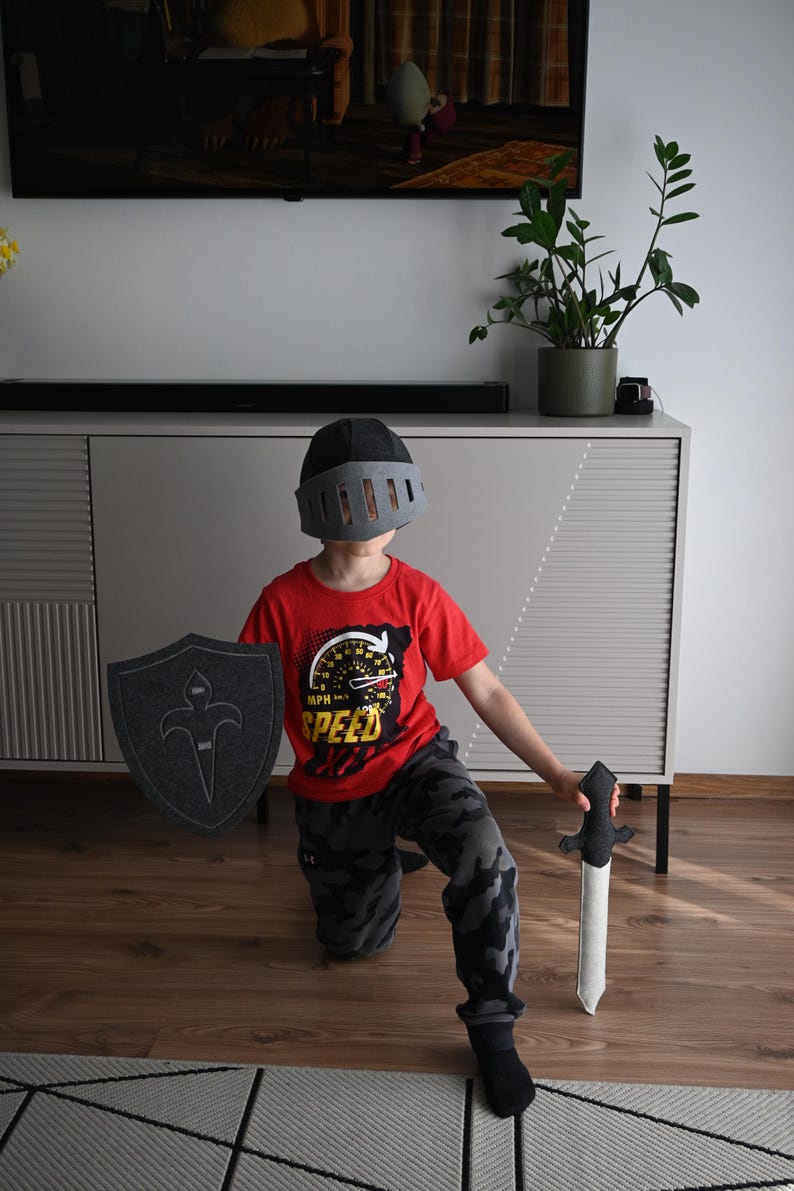 May include: A child wearing a gray helmet, red shirt with a speed graphic, and camouflage pants, holding a gray sword and shield.