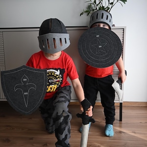 Op de afbeelding: Twee kinderen dragen grijze vilten helmen en houden grijze vilten schilden en zwaarden vast. Een kind draagt een rood shirt en een zwarte broek, het andere kind draagt een rood shirt en een camouflagebroek.
