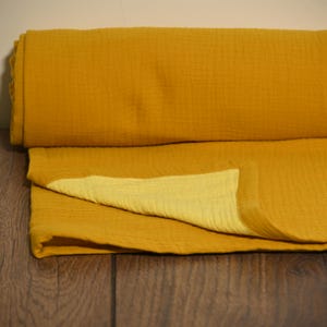 May include: Mustard yellow muslin fabric, folded and rolled. The fabric has a textured, crinkled appearance. The folded portion reveals a lighter yellow hue on the underside. The fabric is likely used for blankets, swaddles, or clothing.