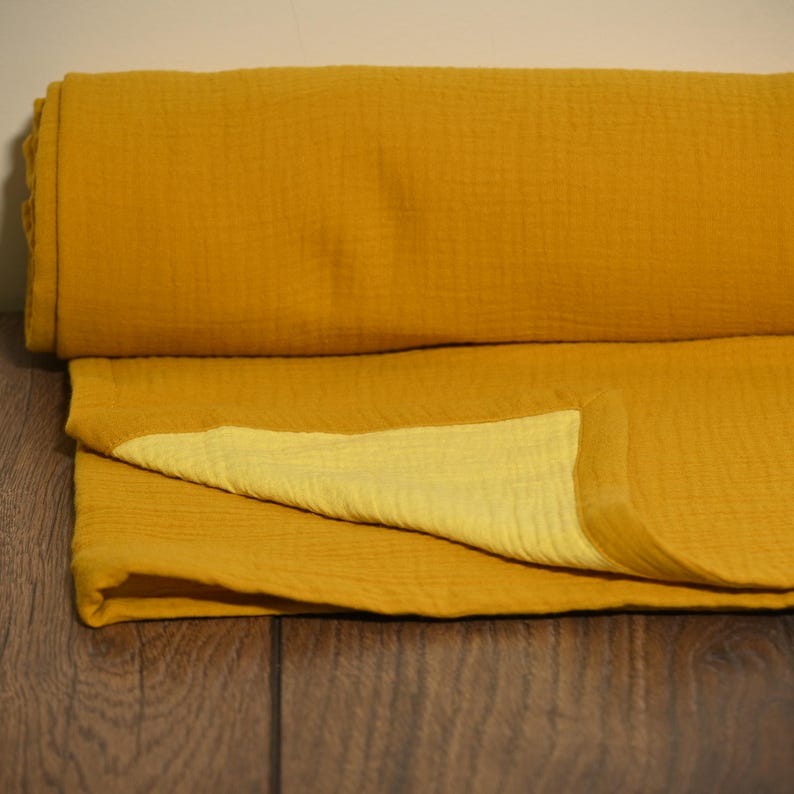 May include: Mustard yellow muslin fabric, folded and rolled. The fabric has a textured, crinkled appearance. The folded portion reveals a lighter yellow hue on the underside. The fabric is likely used for blankets, swaddles, or clothing.