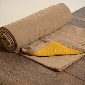 May include: A rolled-up, tan-colored blanket with a yellow border. The blanket is made of a soft, textured fabric and is partially unrolled, revealing the yellow accent. The blanket rests on a wooden surface.