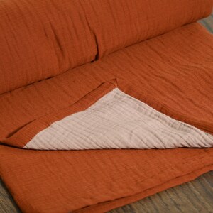 May include: A folded, rust-colored blanket with a textured surface. The blanket is made of a soft material and has a light beige underside. The blanket is folded over, revealing the contrasting color of the underside.