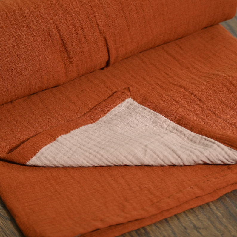 May include: A folded, rust-colored blanket with a textured surface. The blanket is made of a soft material and has a light beige underside. The blanket is folded over, revealing the contrasting color of the underside.