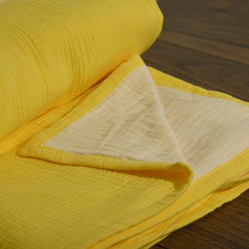 May include: A folded yellow blanket with a cream-colored interior and yellow trim. The blanket is made of a soft, textured fabric. The blanket is partially rolled up, revealing the layers and texture of the material.