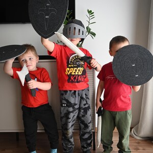 May include: Three children playing knights with toy swords and shields. One child is wearing a gray helmet and holding a gray shield with a silver sword. Another child is holding a gray shield with a red sword. The third child is holding a gray shield with a black sword. All three children are wearing red shirts and black pants.