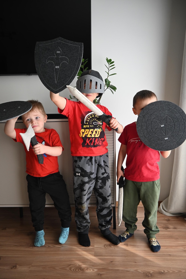 May include: Three children playing knights with toy swords and shields. One child is wearing a gray helmet and holding a gray shield with a silver sword. Another child is holding a gray shield with a red sword. The third child is holding a gray shield with a black sword. All three children are wearing red shirts and black pants.