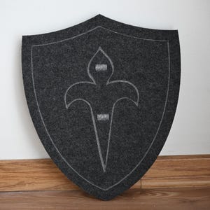 May include: A gray felt shield with a white stylized fleur-de-lis design.