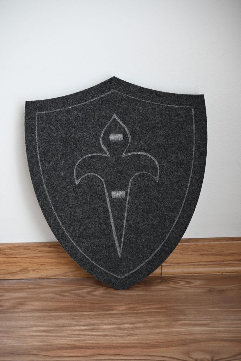 May include: A gray felt shield with a white stylized fleur-de-lis design.