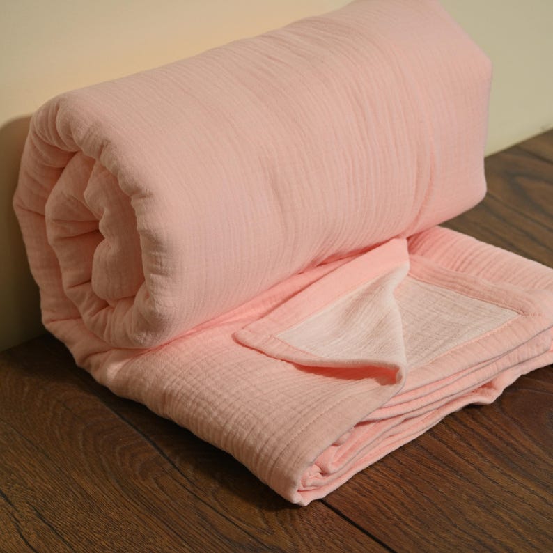 May include: A rolled-up, light pink blanket with a textured, crinkled appearance. The blanket is partially unrolled, revealing a lighter pink interior and a contrasting border. The soft, folded fabric suggests comfort and warmth.
