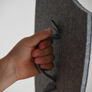 May include: A gray felt shield with a cut-out handle. The shield is held by a hand.