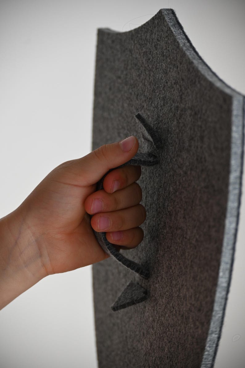 May include: A gray felt shield with a cut-out handle. The shield is held by a hand.