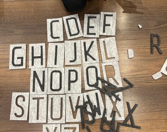 May include: A set of felt alphabet letters in gray with black lettering. The letters are arranged in alphabetical order on a wooden floor.