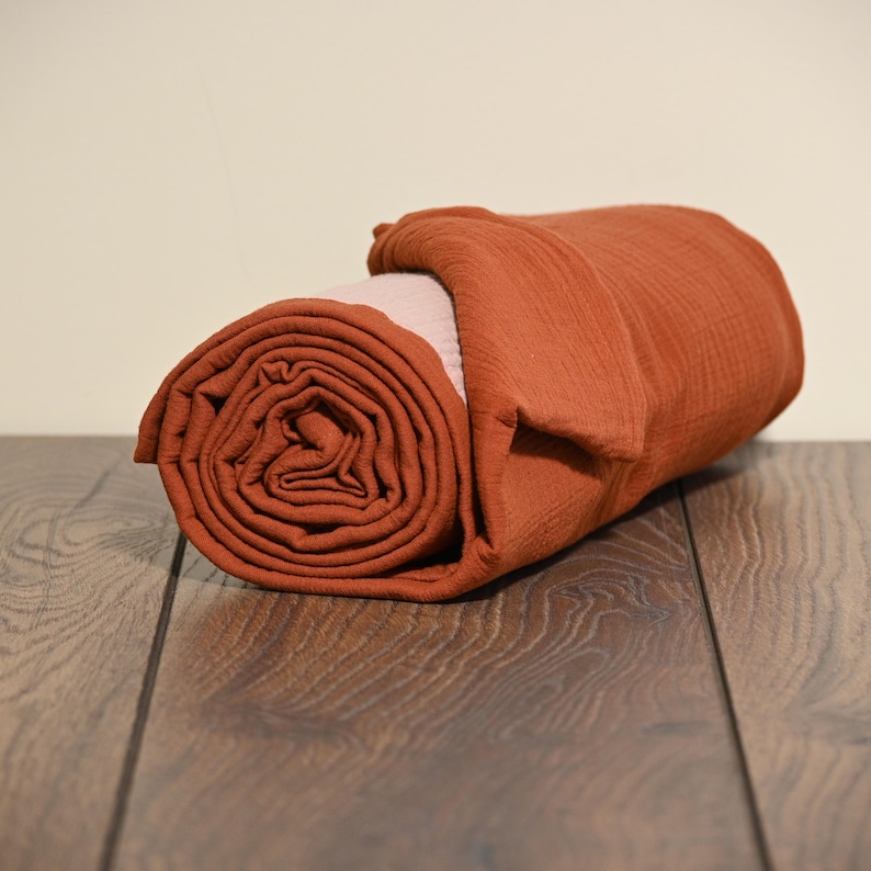 May include: Rolled terracotta-colored blanket with a light pink interior. The blanket is made of a textured fabric and is neatly rolled up, showcasing its layered design. The blanket is resting on a dark wood surface.