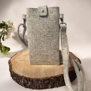 May include: A light gray felt crossbody phone pouch with a rectangular shape and a single silver-toned metal snap button closure at the top center. The bag features a matching gray felt shoulder strap attached with silver-toned metal swivel clips.