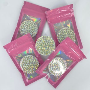 Puede incluir: Se muestran cinco agarres redondos para teléfono con diamantes de imitación iridiscentes. Cada agarre está dentro de una bolsa rosa resellable. Los agarres están cubiertos de pequeños diamantes de imitación brillantes, creando un efecto brillante. El fondo es blanco liso.