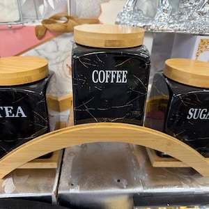 May include: A trio of black ceramic storage jars with bamboo lids, labelled "TEA", "COFFEE", and "SUGAR". The jars feature a gold marble effect and are displayed on a bamboo arched stand. A stylish kitchen storage solution.