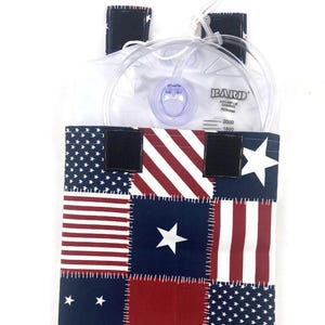 May include: A rectangular fabric cover featuring a patchwork design of red, white, and blue stars and stripes. The cover has black hook-and-loop fasteners at the top and holds a clear plastic Bard urinary drainage bag with visible tubing and markings.