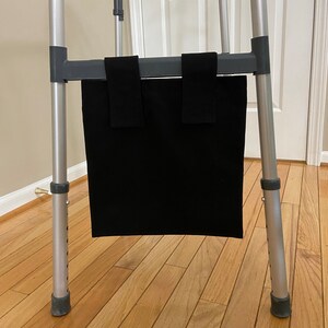 May include: A black walker bag attached to a silver and gray walker. The bag is rectangular and hangs from the walker's frame with two black straps. The walker has adjustable legs and rubber tips.
