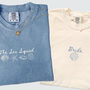 Könnte beinhalten: Ein blaues T-Shirt mit weißer Stickerei, die The Sea Squad über vier Muschelsymbolen zeigt, und ein elfenbeinfarbenes T-Shirt mit blauer Stickerei, die Bride über einem einzelnen Muschelsymbol zeigt, beide mit Comfort Colors Etiketten.
