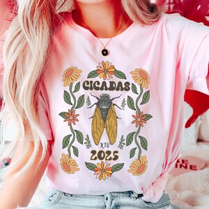 May include: A pink t-shirt with a graphic of a cicada surrounded by flowers and the text "CICADAS XIV 2025".