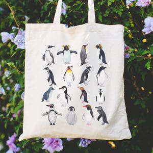 May include: A cream-colored tote bag featuring a collection of watercolor penguin illustrations. The penguins vary in size and pose, with black, white, gray, and yellow coloring. The bag is held up against a backdrop of green foliage and purple flowers.