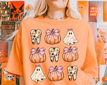May include: Orange t-shirt with a Halloween themed design. The design features pumpkins, ghosts, and teeth with a pink bow on each pumpkin. The teeth are decorated with a leopard print pattern.
