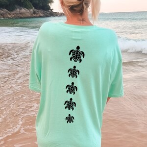 May include: A light green t-shirt with a series of five black turtle silhouettes printed vertically down the back. The shirt is a solid color and appears to be made of a soft material. The image is taken outdoors near a beach.