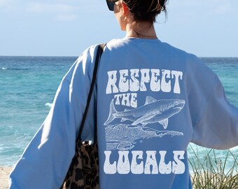 May include: A light blue sweatshirt with a white graphic print of a shark and the text "Respect the Locals" in a retro font.