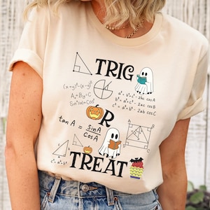 Puede incluir: Una camiseta beige con un diseño gráfico que presenta una calabaza, fantasmas y ecuaciones matemáticas. El texto en la camiseta dice "TRIG OR TREAT".