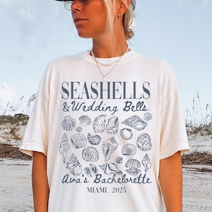 May include: A white t-shirt with a blue graphic of seashells and the text "Seashells & Wedding Bells Ava's Bachelorette Miami, 2025".