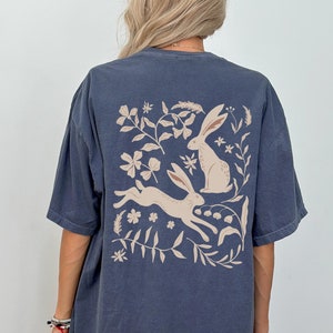 Pu&ograve; includere: Una maglietta oversize blu scuro con una grafica beige di due conigli e disegni floreali sul retro.