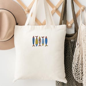 May include: A white canvas tote bag with a design of five colorful fish on the front. The bag is hanging on a wooden rack with a brown hat and a beige mesh bag.