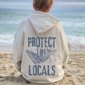 Może przedstawiać: Kremowa bluza z kapturem z niebieską grafiką wieloryba i napisem "PROTECT THE LOCALS". Projekt przedstawia szczeg&oacute;łową ilustrację wieloryba wyskakującego z wody, z postarzaną czcionką. Bluza noszona jest na piaszczystej plaży z oceanem w tle.