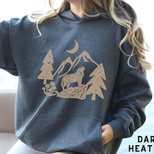 Puede incluir: Sudadera con capucha de color gris oscuro jaspeado con un estampado gr&aacute;fico de un lobo aullando a la luna en un paisaje de monta&ntilde;a. El gr&aacute;fico est&aacute; en un estilo vintage y desgastado.